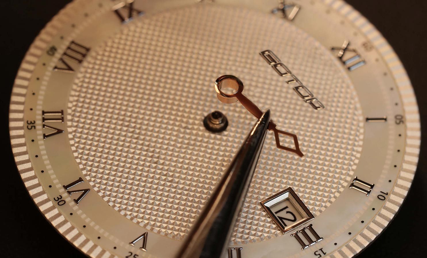 Hands being fitted to a gold Roman numeral dial with tweezers.